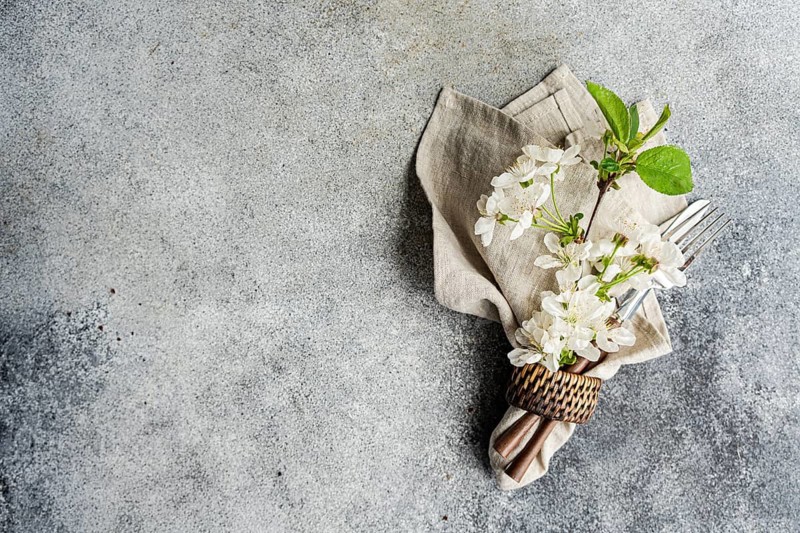 An elegant table setting with a folded linen napkin, delicate white flowers, and silver fork and knife on a textured stone surface creates a sophisticated dining atmosphere