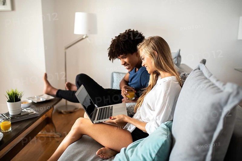 A couple sits comfortably on a couch in their living room, sharing a laptop. Natural light enhances the room's cozy ambiance, creating a relaxed and intimate atmosphere