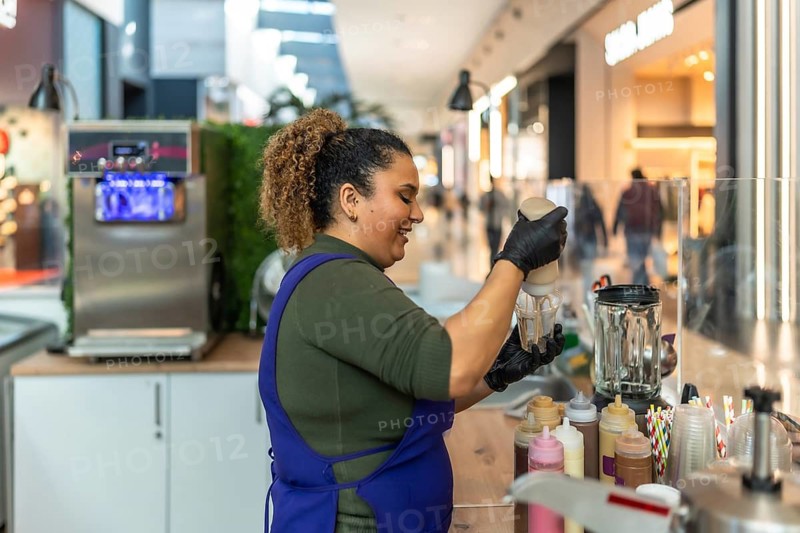 Smiling woman wearing an apron and gloves pouring topping into a cup, preparing a refreshing milkshake for a customer at a small business food stand in a busy shopping mall