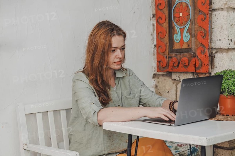 Woman in her 40s works on a laptop at an outdoor table, appearing serious and concentrated. She is engaged in freelance tasks or online study in a serene setting