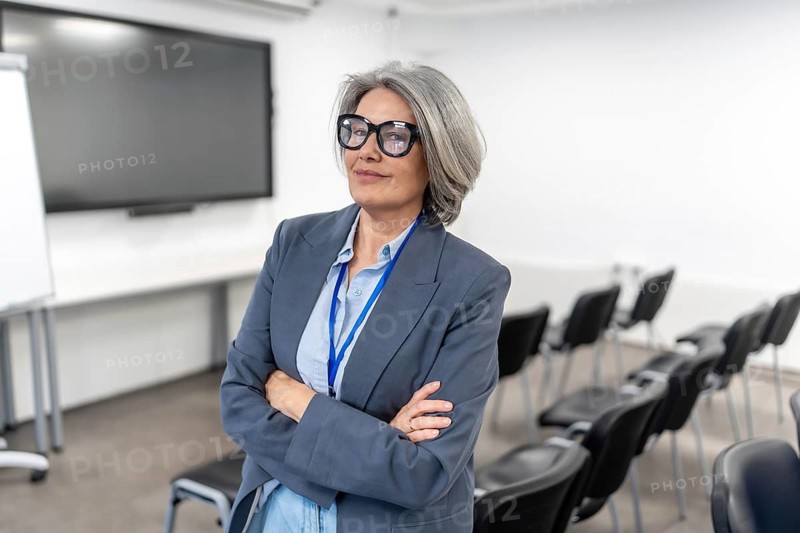 Mature business professional woman with grey hair and glasses standing confidently with arms crossed in a modern empty conference room, preparing for a corporate event or educational presentation