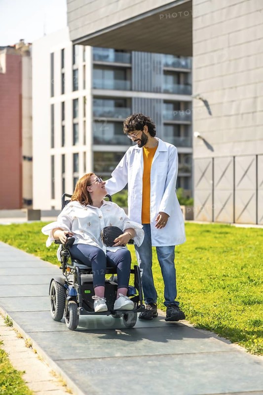 Doctor assisting a female patient in an electric wheelchair, strolling together outdoors in a park, showcasing healthcare and mobility support
