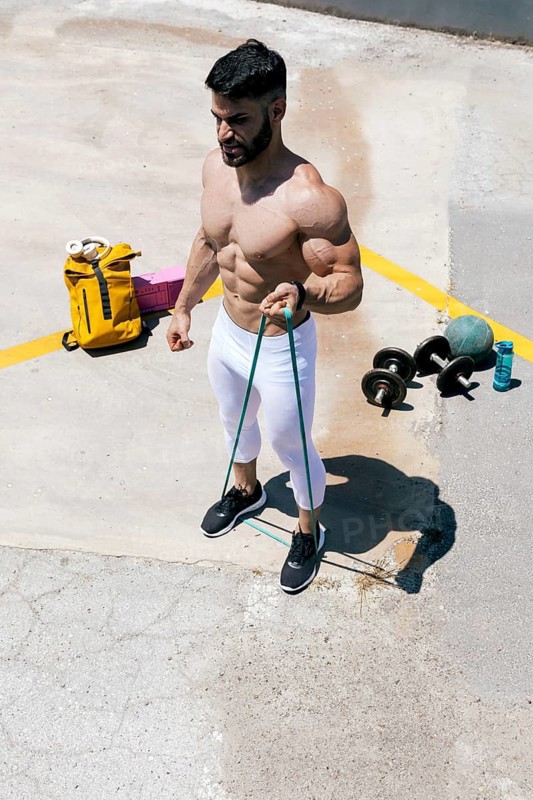 A muscular man exercises outdoors on a sunny day, using resistance bands. Fitness equipment, including weights and a water bottle, is scattered on the concrete surface