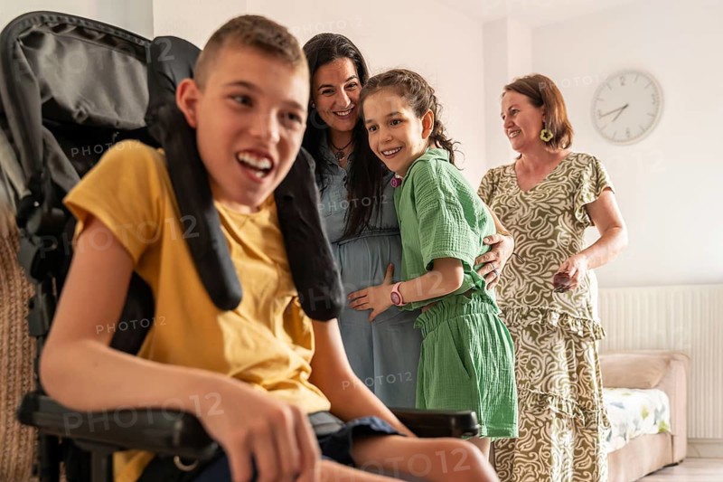 A joyful family shares a moment at home, highlighting love and diversity. A disabled girl with a tracheostomy and a boy with cerebral palsy smile brightly with their family