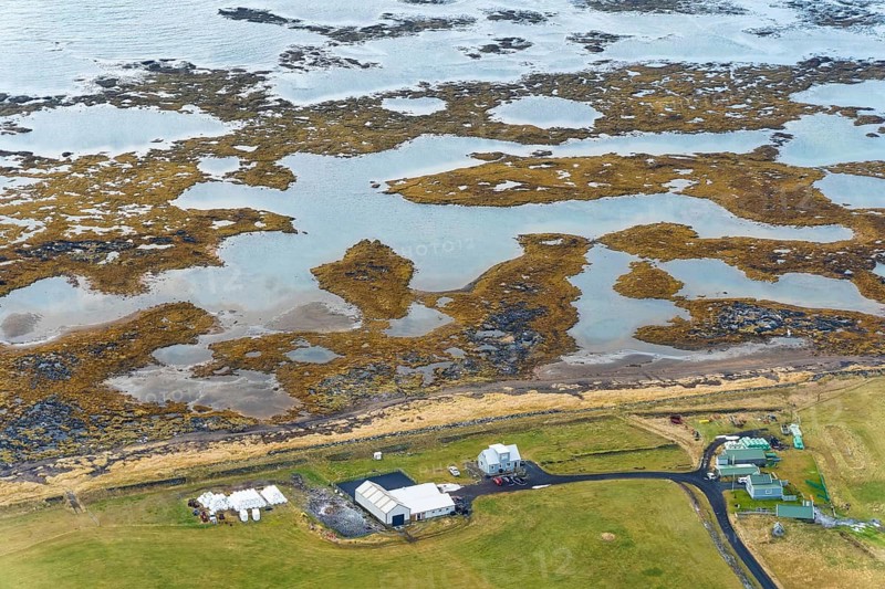 Aerial view of a small village in Iceland, near Reikiavik, featuring scattered houses and natural tidal wetlands along the rugged coastline
