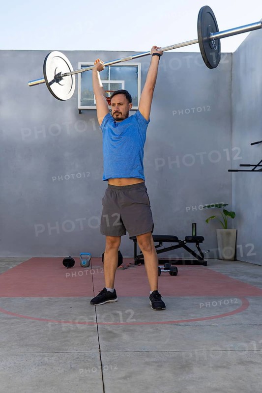 Focused sportsman lifting a barbell overhead, engaging in strength training exercises within an outdoor home gym, showcasing dedication to fitness and a healthy lifestyle