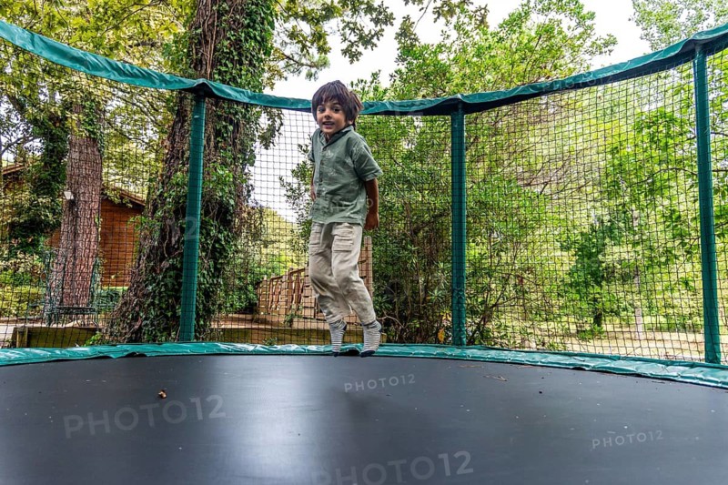 Young child experiencing joy and freedom while bouncing on an outdoor trampoline with a safety net, spending active leisure time in a green natural setting in messanges, landes, france
