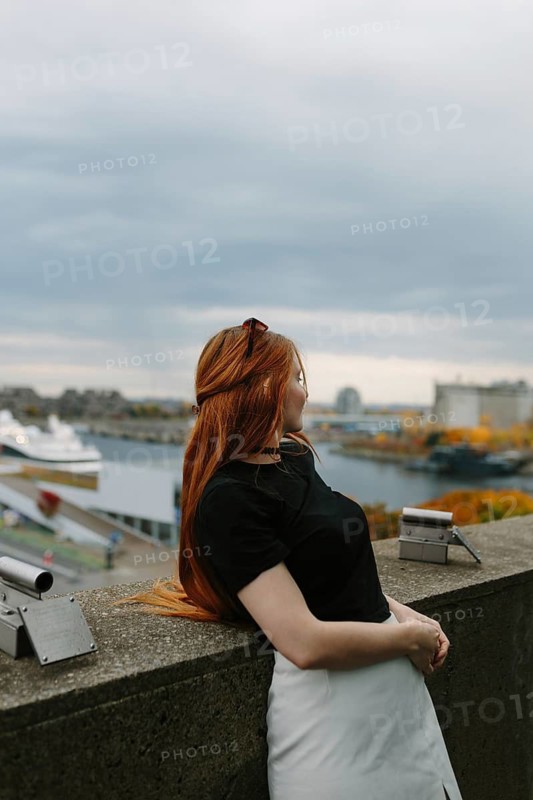A red-haired woman in a black top and white skirt leans on a stone wall, enjoying the scenic view of Montreal's harbor. Autumn leaves and a cloudy sky complete the urban backdrop