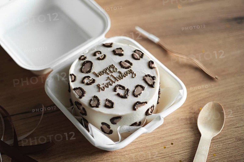 South Korean bento birthday cake featuring Happy Birthday written with chocolate decorations, presented in a white takeaway box, alongside a wooden spoon and matte ribbon