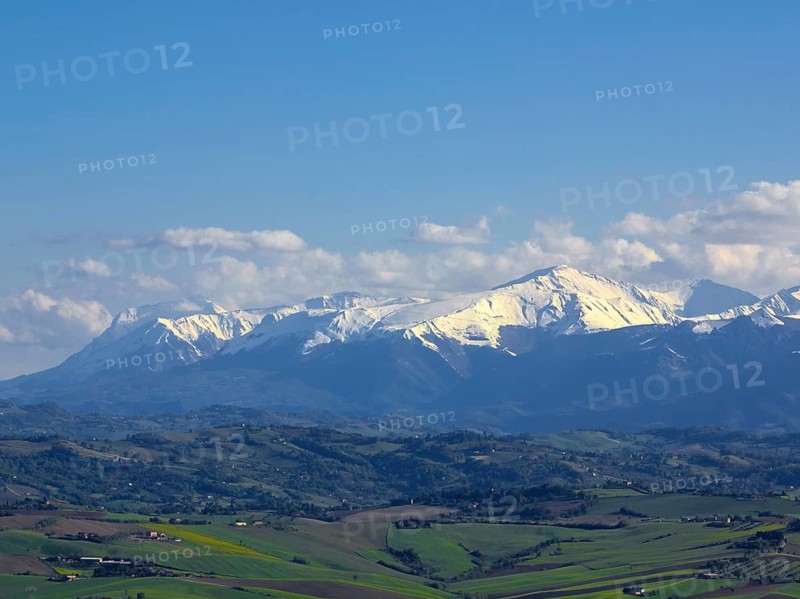 A breathtaking view of snow-capped mountains under a clear blue sky over lush green Italian countryside, illustrating the natural beauty and tranquility of Italy's landscapes