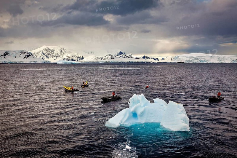 An impressive, cold sea panorama with an iceberg and several small boats under a cloudy sky, tourists in kayaks rowing across the sea in Antarctica