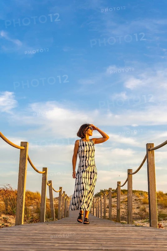 Woman in a summer dress and sunglasses walking along a wooden boardwalk through sand dunes, shielding her eyes from the sun and enjoying a relaxing holiday at the beach during sunset