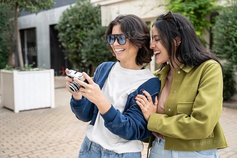 A joyful lesbian couple laughing while viewing photos on a camera. They are enjoying each other's company in a sunny outdoor setting, showcasing love and happiness