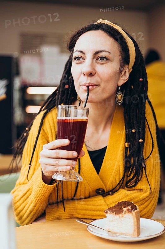 A woman in a yellow sweater and dreadlocks enjoys a drink through a straw, sitting at a table with a delicious slice of cake in a warm, inviting cafe atmosphere