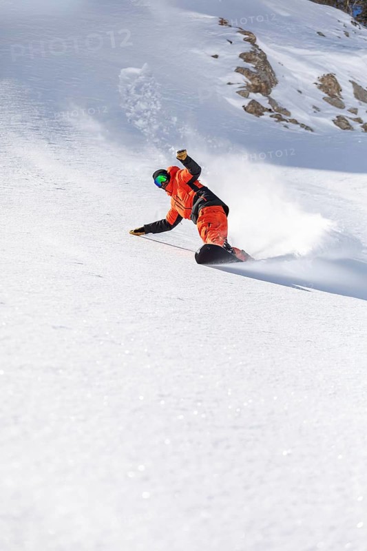 Snowboarder in bright orange gear skillfully carves down a snowy mountain slope. Captured in action, the scene highlights dynamic motion and winter sports excitement