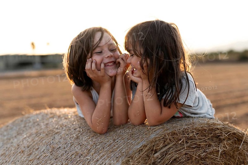 Two young children share a joyful moment on a bale of hay in a sunny rural field. Their laughter and carefree expressions capture the essence of childlike happiness