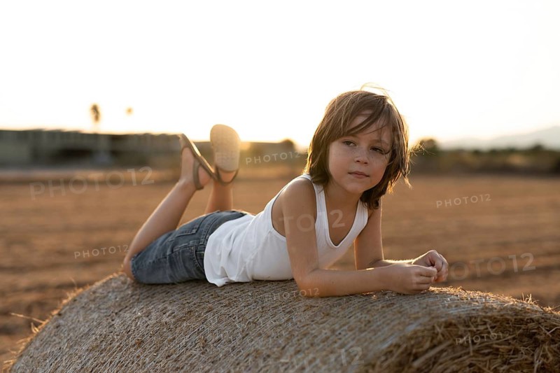 Child in casual attire lies on a hay bale, surrounded by a scenic field at sunset. The soft lighting and relaxed pose create a serene and nostalgic atmosphere
