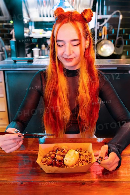 A woman with long, vibrant hair savors a dish with chopsticks at an Asian food market She smiles, sitting at a wooden table in a lively and colorful market setting