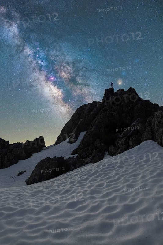 A lone figure stands on a snowy Icelandic peak beneath a stunning Milky Way sky The scene captures the serene beauty of a clear northern night in Iceland