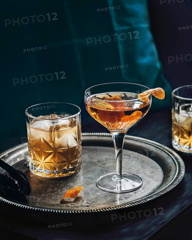 A sophisticated whiskey and orange cocktail sits elegantly on an ornate silver tray, accompanied by two glasses. The cocktail is garnished with a zesty orange peel