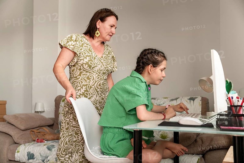 A caring family moment showing a mother assisting her disabled daughter, who has a tracheostomy, while engaging with the computer