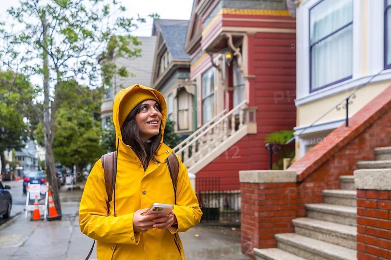 Woman in yellow raincoat and beanie walking a san francisco street of colorful victorian and edwardian homes, smiling and exploring the historic neighborhood with phone in hand