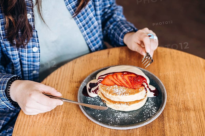 A close up shot of a woman enjoying fluffy japanese pancakes topped with fresh strawberries and syrup. The setting is cozy, with a wooden table adding warmth to the scene