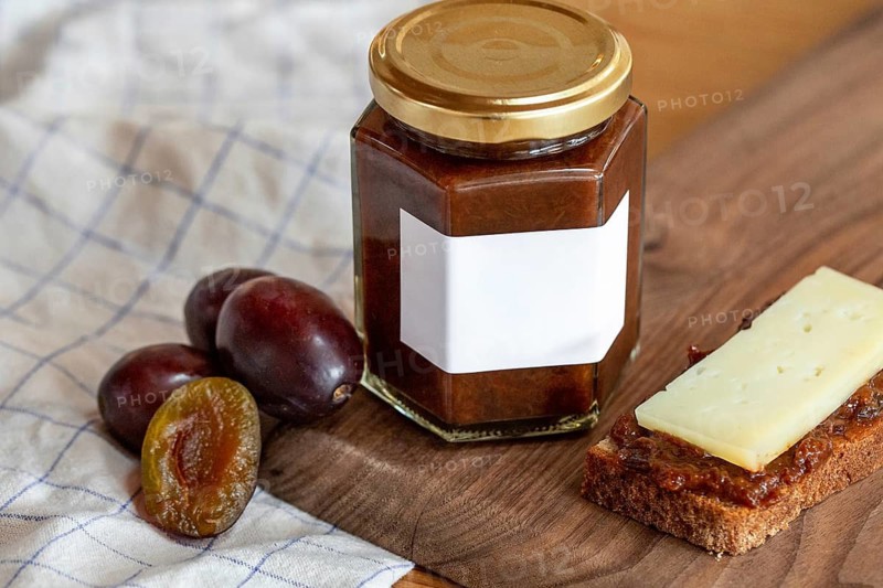 A hexagonal jar of plum compote with a blank label is displayed on a wooden surface. Fresh plums and a slice of bread with compote and cheese accompany the scene for a rustic touch