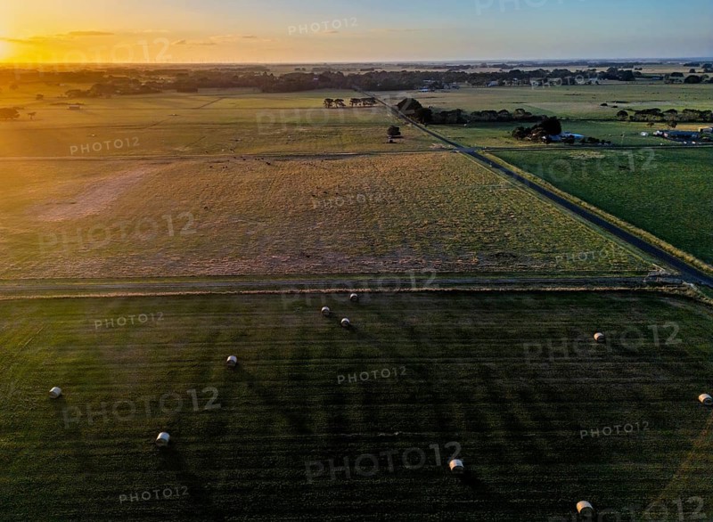 Aerial view of a sprawling countryside landscape with lush green fields and hay bales scattered around, bathed in the golden glow of the setting sun in Victoria, Australia