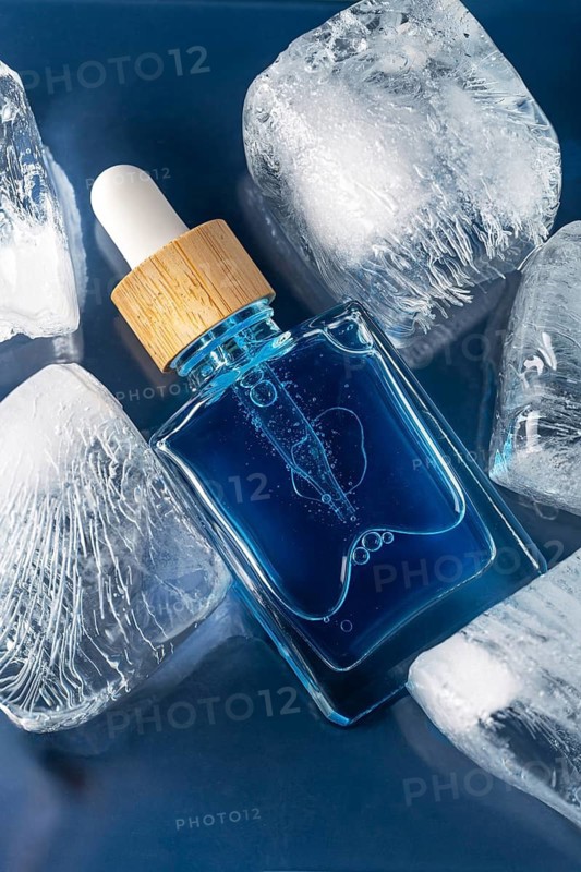 Top view of a face care serum or oil for men showcased in a clear dropper bottle, emphasizing its cooling effect. The macro shot highlights a pipette amidst glistening ice cubes