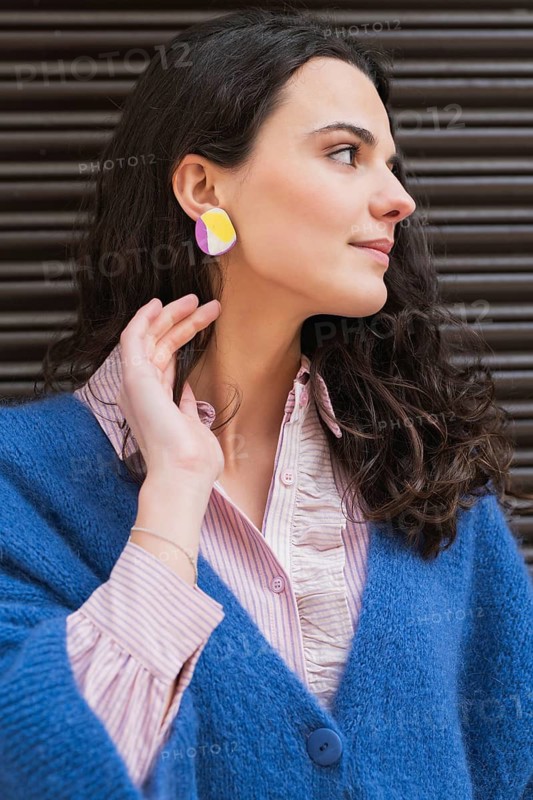 Portrait of a woman wearing a vibrant blue sweater and colorful geometric earrings. She poses gracefully against a dark striped background, showcasing her stylish look