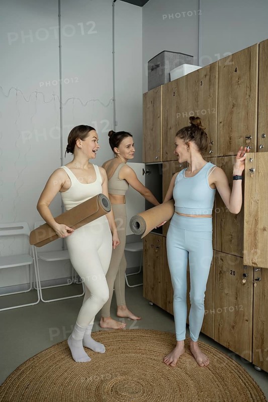 Three women in active wear chat animatedly in a modern locker room before a yoga class, holding mats and preparing for the session in a relaxed environment