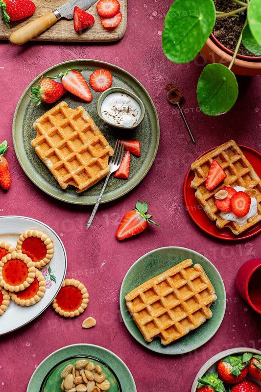 Rustic waffles served with fresh strawberries and yoghurt on colorful plates. A romantic, homemade dessert setup featuring sweet treats and almonds against a vibrant backdrop