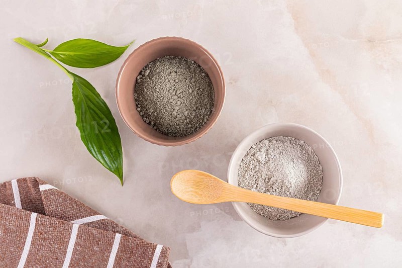 Top view of two clay bowls filled with fine cosmetic clay powder, accompanied by a fresh green leaf and wooden spoon, set on a marble background for skincare routines