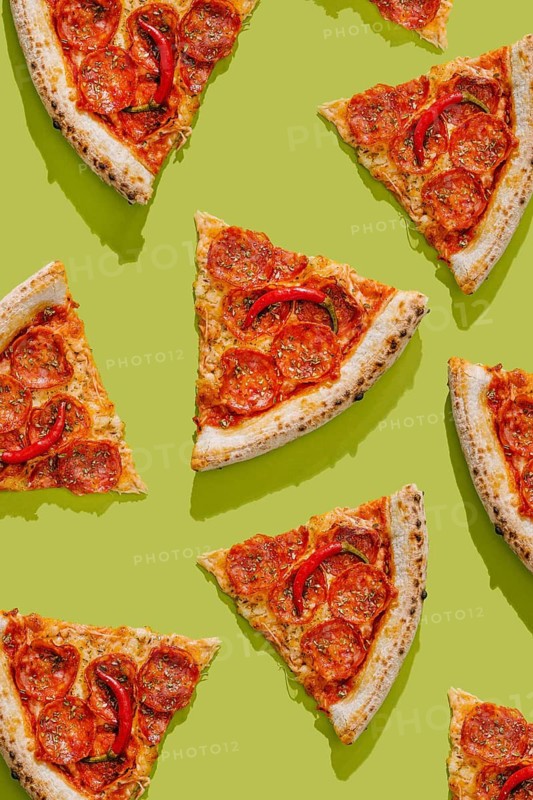 Top view of a pizza sliced into pieces, topped with tomato sauce, pepperoni, mozzarella cheese, hot red peppers, and sprinkled with herbs. Bright green background enhances the vibrant colors