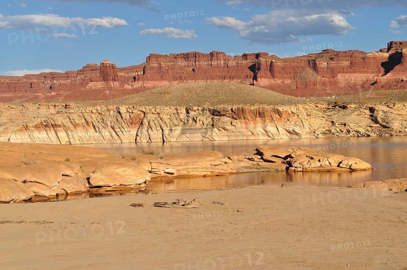 Red rocks tower over a desert-like landscape with calm water under blue skies, Lake Powell, Arizona, USA