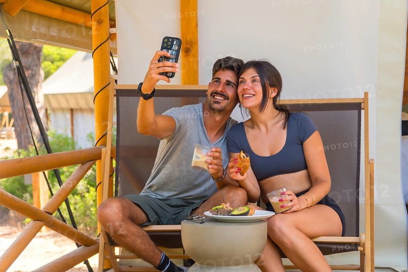 Young couple smiling and taking a selfie with smartphone while having breakfast in a glamping tent, enjoying a relaxing vacation surrounded by nature