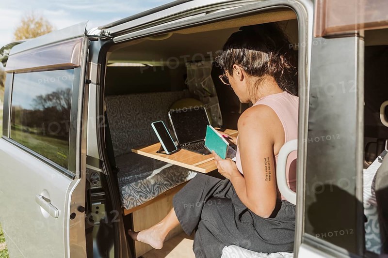 A woman is working inside a van, surrounded by the vibrant autumn foliage of New Zealand. She holds a notebook while using a laptop, embodying a nomadic lifestyle