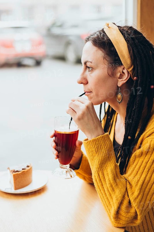 A woman with a yellow headband and warm sweater enjoys a drink through a straw by the window of a cafe Beside her, a slice of cake awaits on the table