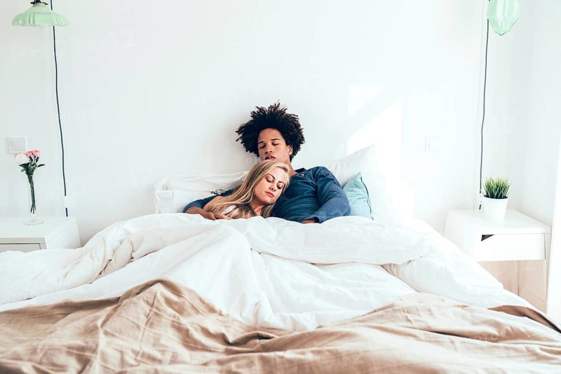 A couple sleeps peacefully on a bed in a minimalist bedroom with white linens and simple decor. Natural light enhances the serene and tranquil atmosphere