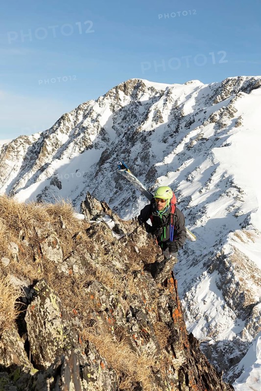 A ski tourer in colorful gear climbs a rugged, snowy mountain ridge under a bright blue sky. The scene captures the thrill and challenge of alpine adventure