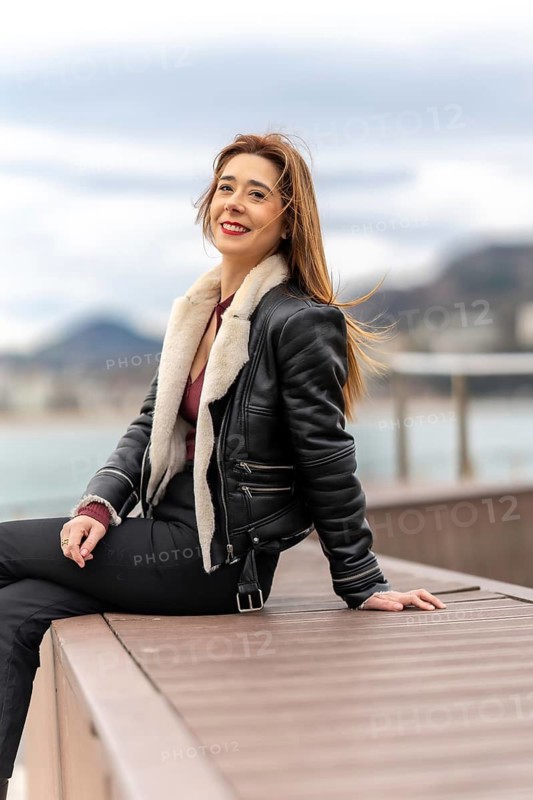 Cheerful woman enjoying a relaxing moment by the sea, wearing a stylish leather jacket and exuding confidence and happiness