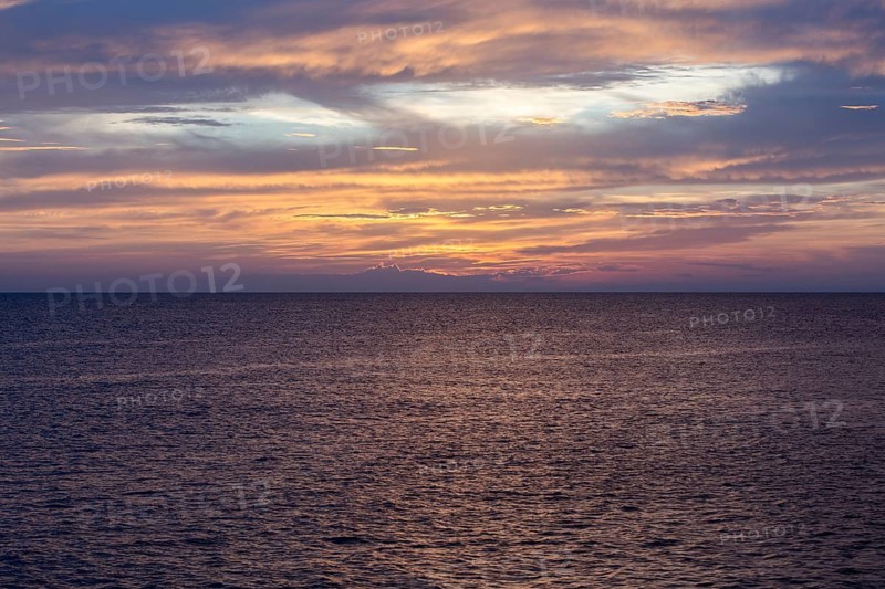 Serene sea scene at sunset with vibrant orange, pink, and purple hues reflecting on the water. The vast sky is painted with mesmerizing clouds, creating a tranquil atmosphere
