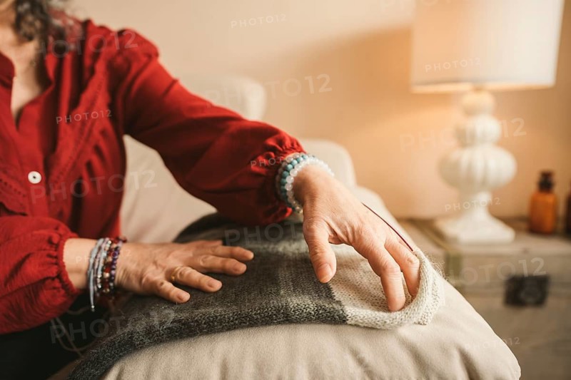 Close-up of a woman's hands adorned with elegant bracelets, touching a cozy, knitted fabric. A warm, inviting home atmosphere is accentuated by a softly lit lamp