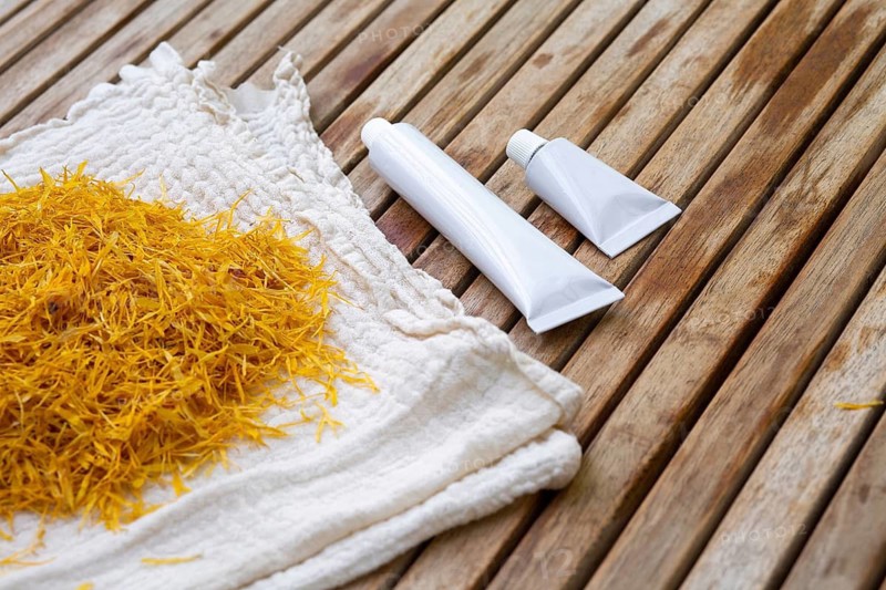 Marigold petals on a cloth beside white cosmetic tubes of marigold petals cream on a wooden surface. The image portrays a natural, herbal approach to skincare and beauty products