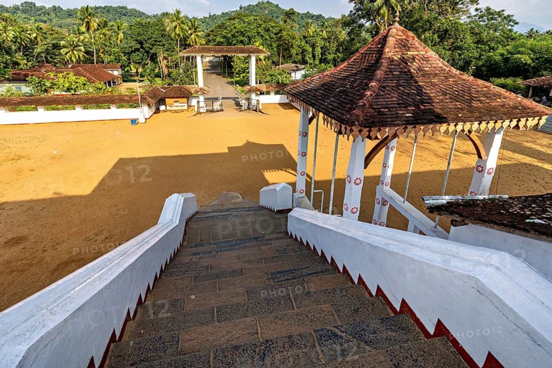 View from a stone staircase of an open sandy square with a traditional pavilion and tropical vegetation, The Sri Sumana Saman Devalaya Temple near Ratnapura in Sri Lanka