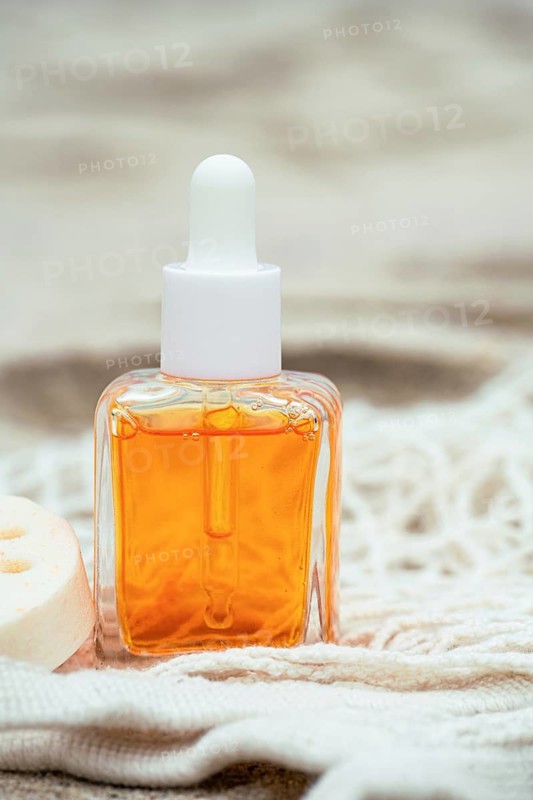 A bottle of hair care oil stands on a sandy textured surface, surrounded by fabric and a loofah. The amber liquid inside contrasts against the natural setting