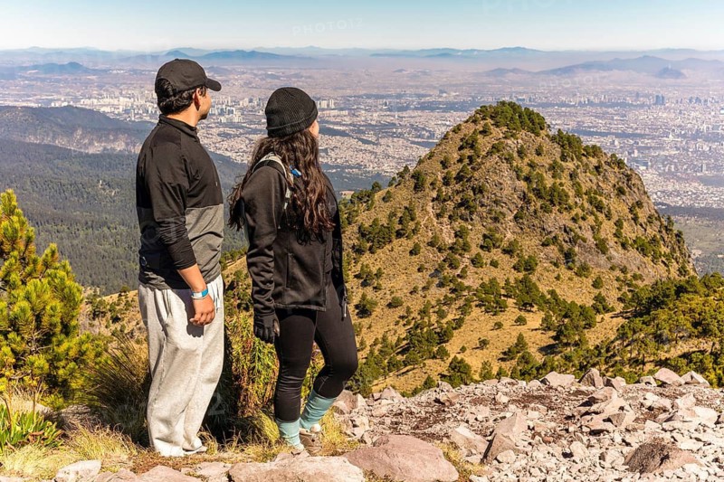 Two friends admire a stunning mountain view from the summit, surrounded by nature They are wearing casual outdoor clothing against a backdrop of distant cityscape