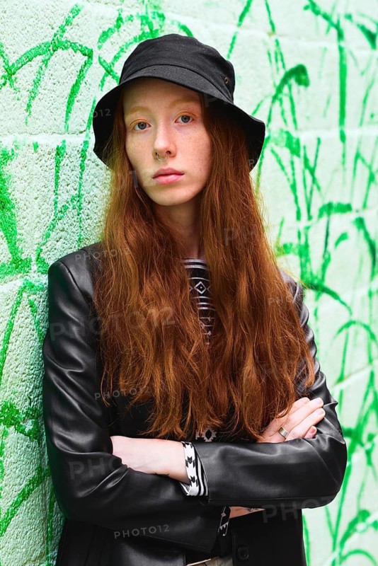 Young woman with long red hair in a stylish black hat and leather jacket poses confidently against a vibrant green graffiti wall backdrop, embodying urban street style
