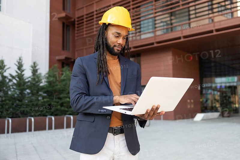 A businessman in a suit and hard hat works on a laptop outside a modern building. The setting combines professional attire with construction elements, highlighting engineering roles
